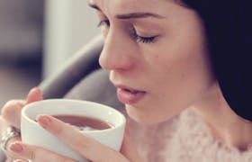 Kvinne sitter med pledd over nakken og drikker varm te for å lindre forkjølelsessymptomer 