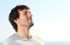Mann med brunt hår står ute i blå himmel og puster med lukkede øyne