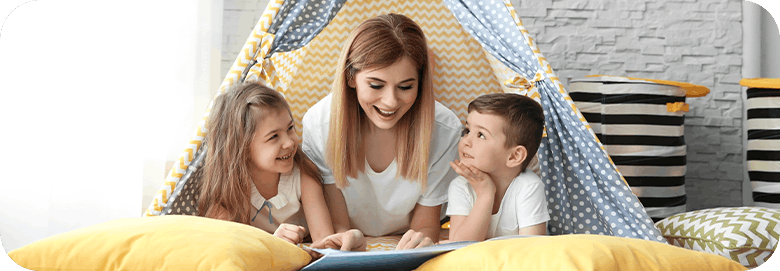 Nanny reading a story book to a young girl and boy laying in a tent at home