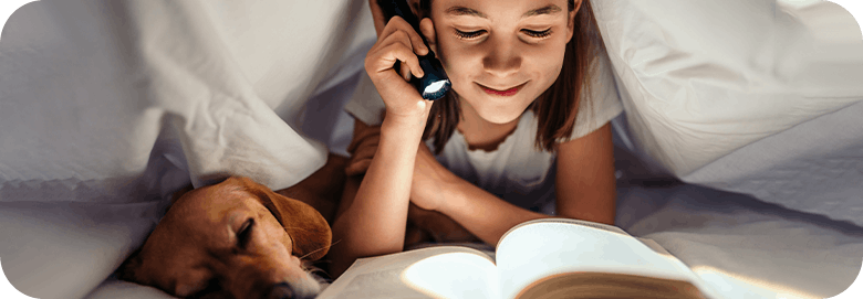 Young girl reading a book under her bed covers with a flash light and her dog beside her