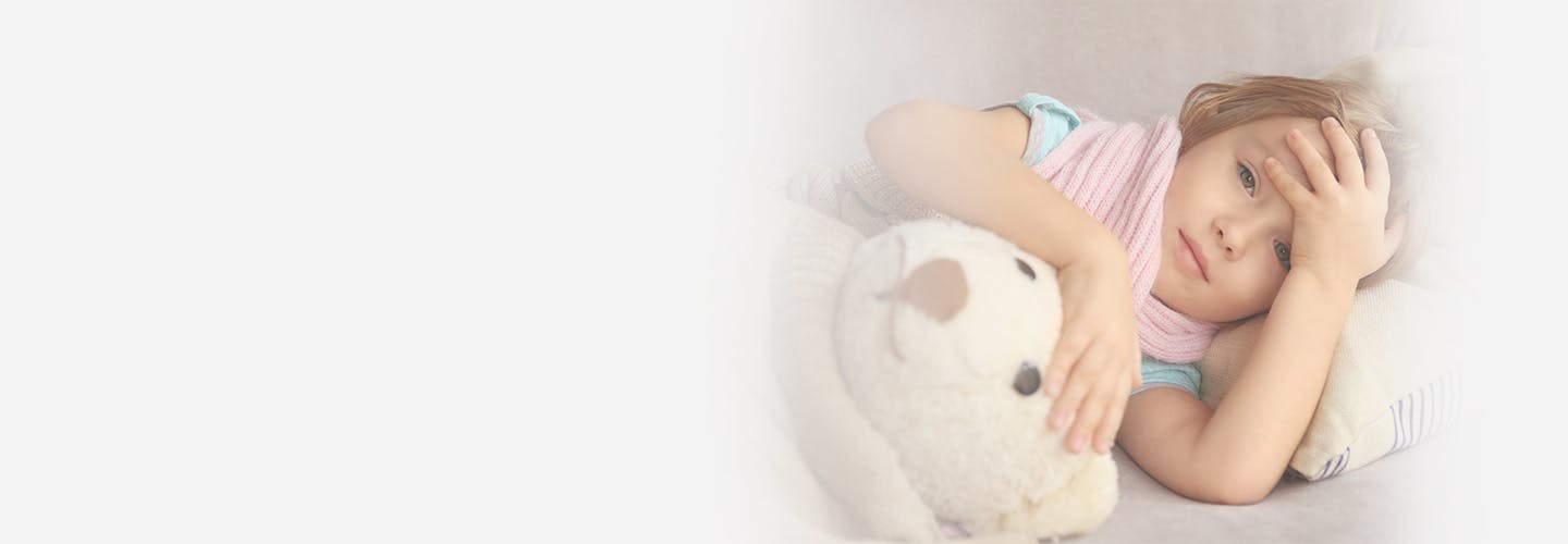 Little girl lying in bed with a teddy bear while holding her head in pain