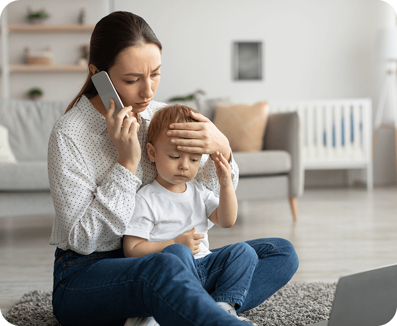Worried woman sitting on floor while holding mobile in one and son's forehead in other hand