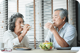 Smiling Asian senior couple sitting at the table drinking glasses of milk and looking at each other