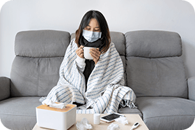 Sick young Asian girl having influenza symptoms wearing medical mask while holding a cup of hot water sitting on sofa