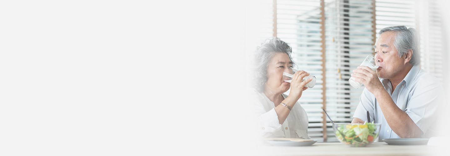 Smiling Asian senior couple sitting at the table drinking glasses of milk and looking at each other