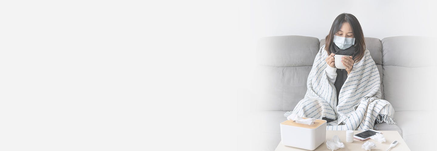 Sick young Asian girl having influenza symptoms wearing medical mask while holding a cup of hot water sitting on sofa