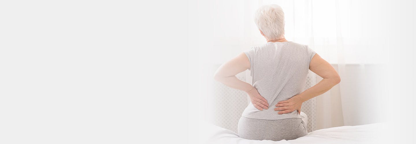 Äldre kvinna sitter på sängkant med ont i ländryggen