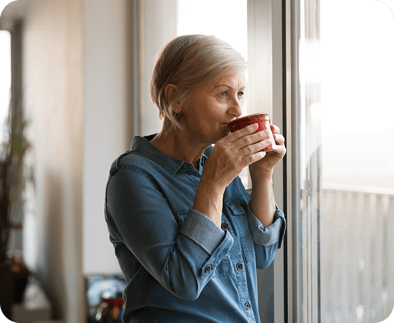 Äldre kvinna dricker en kopp te vid fönster