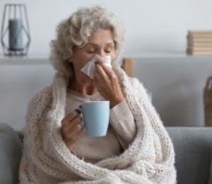 Older adult with nasal congestion, using a tissue and drinking a hot drink. 