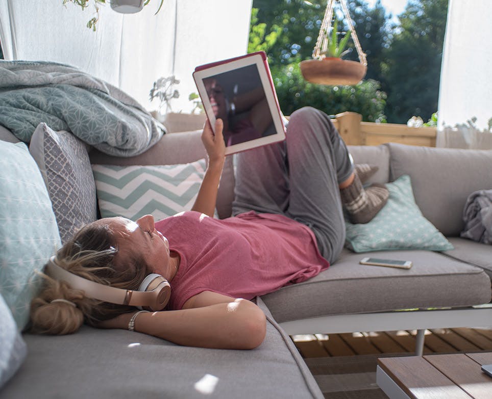 Mujer joven recostada en el sofá escuchando música y viendo su tablet. 