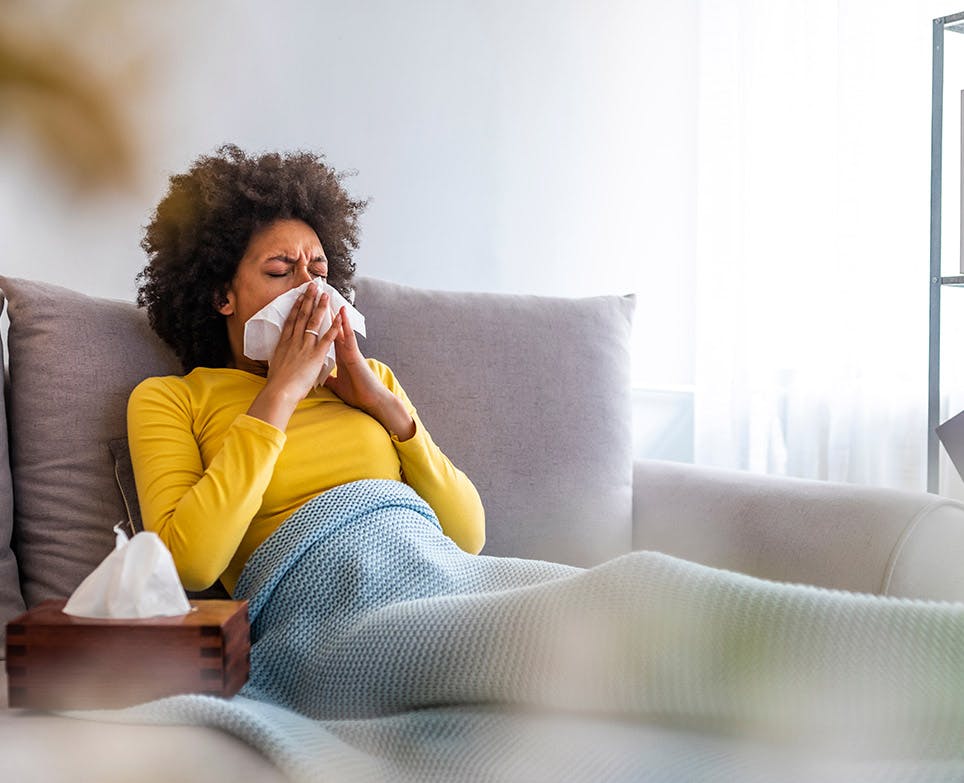 Mujer con congestión nasal, sentada en un sofá, limpiando su nariz. 