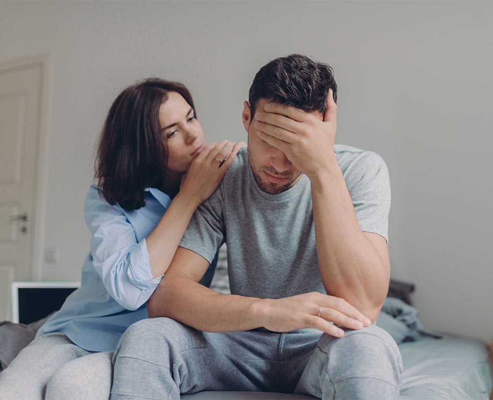 Pareja sentada. El hombre se toca la cabeza en señal de dolor. 