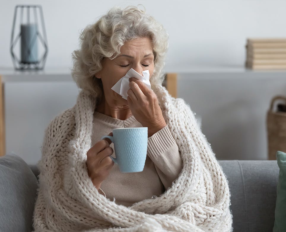 Adulta mayor con congestión nasal, limpiando su nariz y tomando una bebida caliente. 