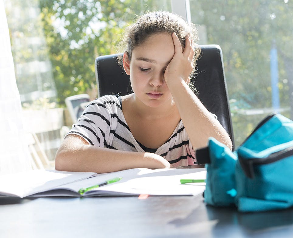 Niña estudiando mientras se toca la cabeza con la mano en señal de dolor. 