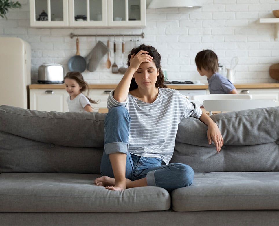 Representación de dolor en una madre joven, sentada en un sofá mientras toca su cabeza y sus hijos juegan. 