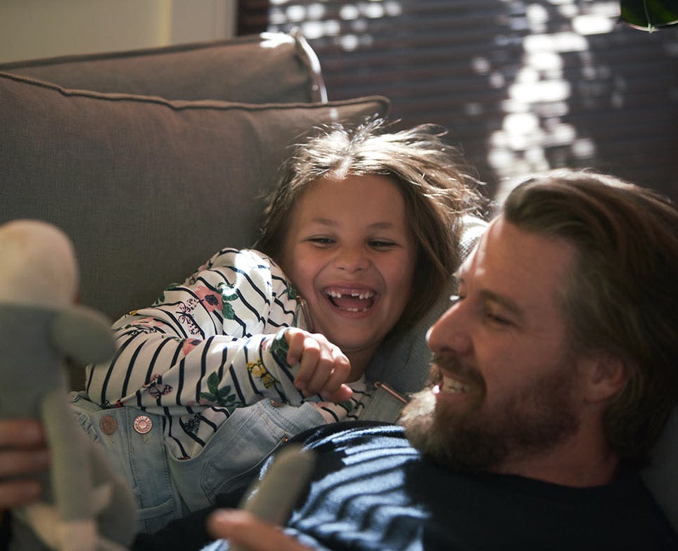 Zolben - Hombre y su hija recostados en un sillón, riendo y jugando con un muñeco