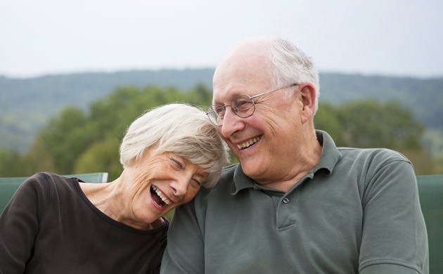 Una pareja de adultos mayores sonríen, sentados en un parque.