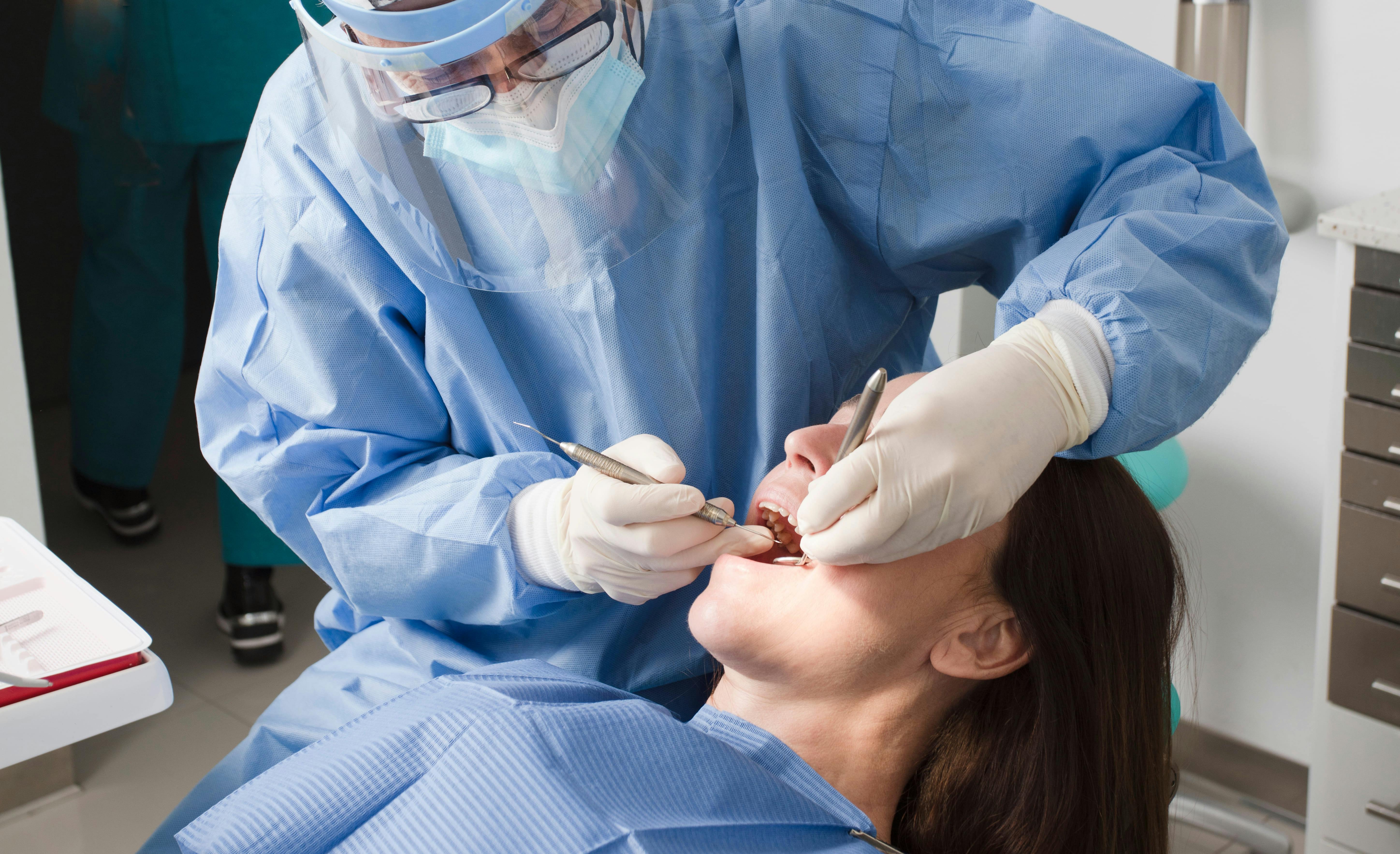 A dentist wearing a mask and face shield works on the teeth of a patient with long hair.