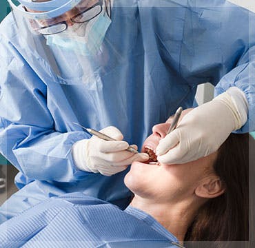 A dentist wearing a mask and face shield works on the teeth of a patient with long hair.