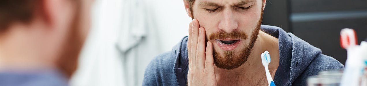 man with sensitive tooth pain holding toothbrush and touching his cheek