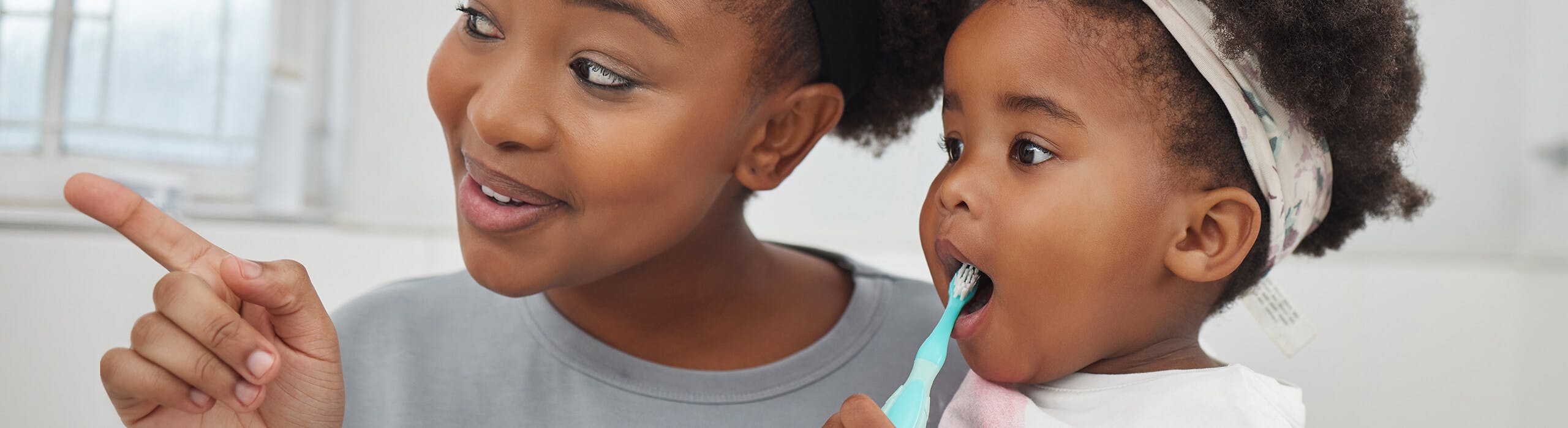 A mother supervising her child as she brushes her teeth with a toddler-friendly toothbrush