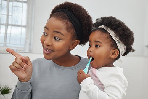 A mother supervising her child as she brushes her teeth with a toddler-friendly toothbrush