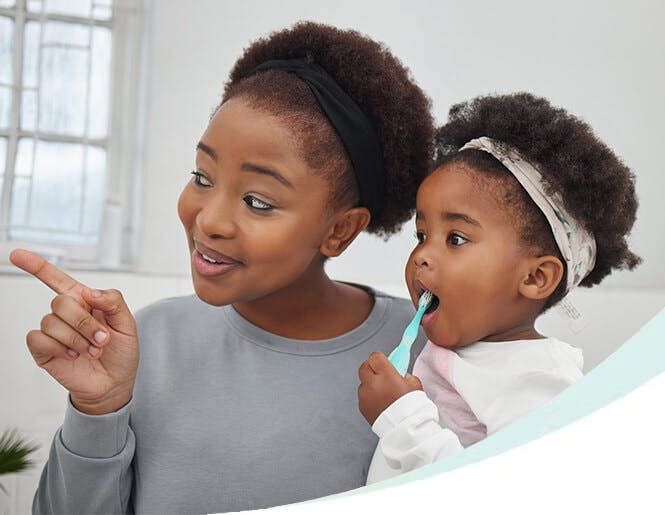 A mother supervising her child as she brushes her teeth with a toddler-friendly toothbrush