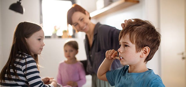 Mother in a bathroom with two little girls and a little boy who is brushing his teeth