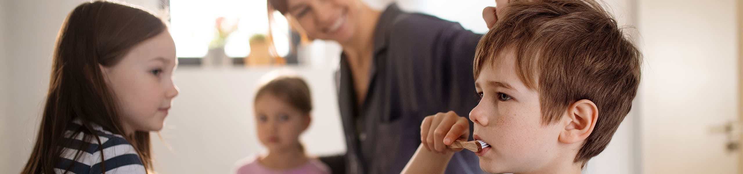 Mother in a bathroom with two little girls and a little boy who is brushing his teeth