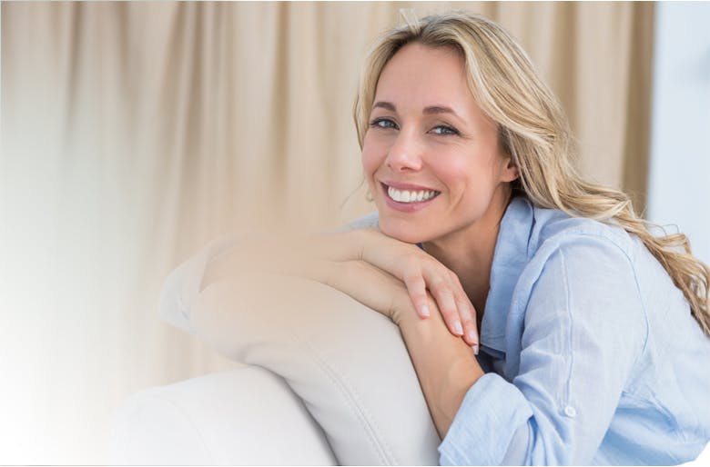 Femme blonde souriante, assise sur un canapé gris, le menton posé sur les mains