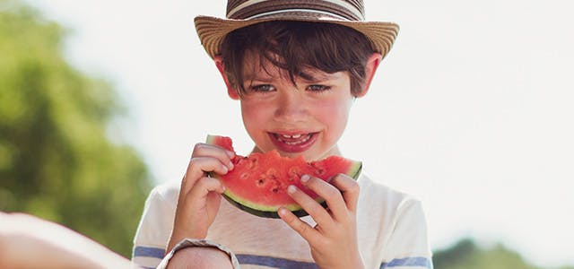 Un garçon mange une tranche de melon d'eau acide tandis que deux fillettes jouent en arrière-plan