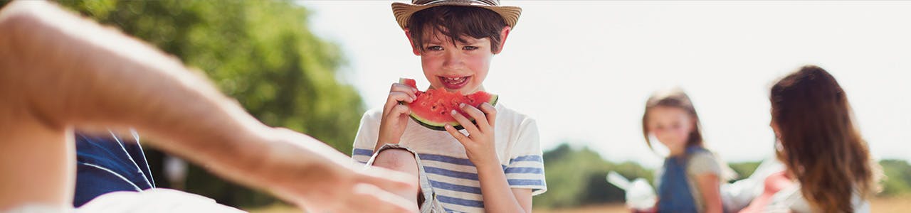 Un garçon mange une tranche de melon d'eau acide tandis que deux fillettes jouent en arrière-plan