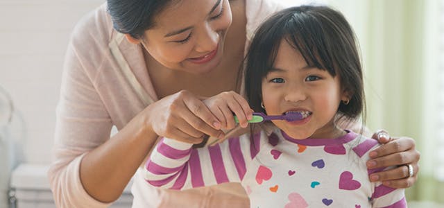 Mom helps her daughter to brush her teeth in front of the mirror with Pronamel® 