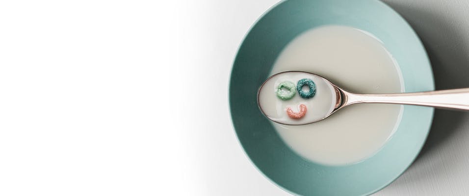 Colourful cereal in the shape of a smiling face surrounded by milk in a spoon on top of a bowl
