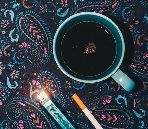 Cup of coffee on a table covered in a paisley tablecloth, sitting beside a cigarette and e-cigarette