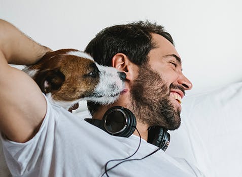 Man laying in his bed, laughing while a dog cuddles into his neck