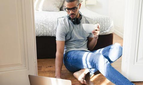 Man sitting in his bedroom, leaning against his bed, drinking a cup of coffee and looking at a laptop