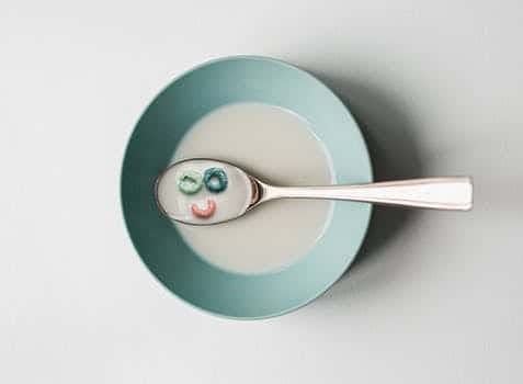 Colourful cereal in the shape of a smiling face surrounded by milk in a spoon on top of a bowl
