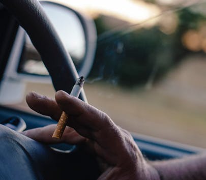 Main d’un homme dans une voiture tenant le volant et une cigarette allumée entre 2 doigts