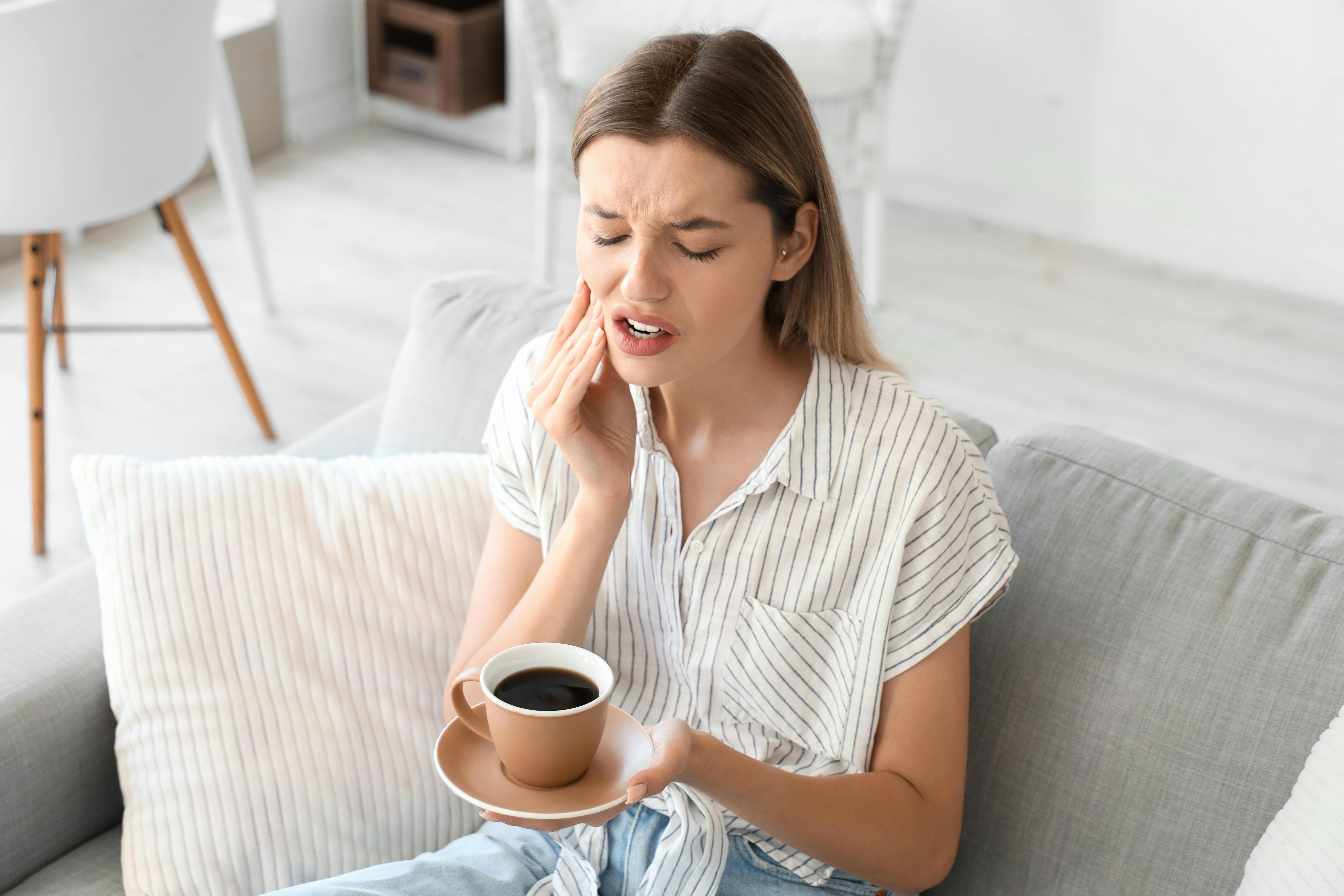 Woman in pain touches her cheek with her right hand and holds a hot cup of black coffee in her left hand