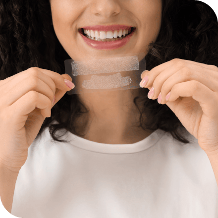 A woman smiling and holding up at-home teeth whitening strips