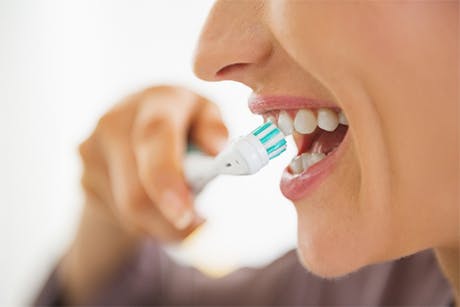 Image of the lower half of a woman's face, smiling as she brushes her teeth with a manual toothbrush