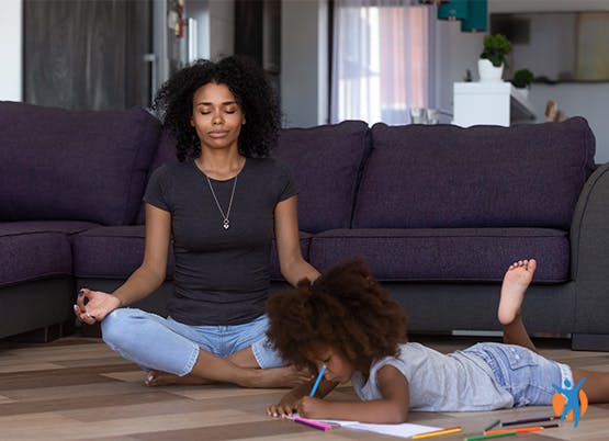 Daughter laying on the living room floor and drawing with coloring pencils while her mother is siting next to her in meditating pose with her eyes closed