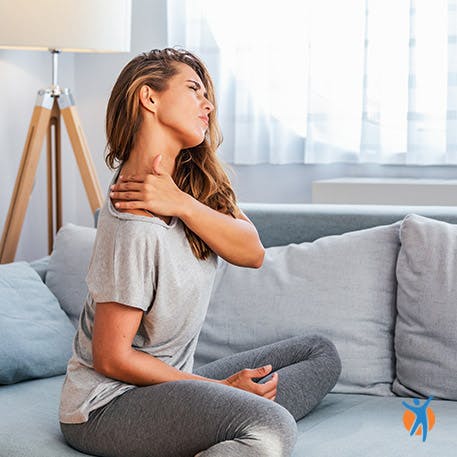 Mulher na sala a massajar o pescoço e com evidência de presença de dor.