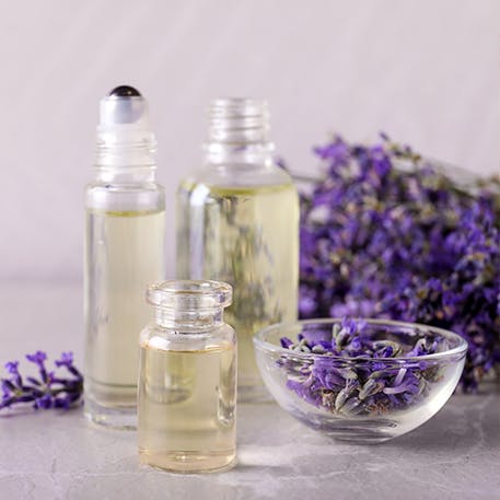 Três garrafas de dimensões diferentes com óleo essencial e algumas flores de lavanda numa taça e um molho de lavanda no fundo