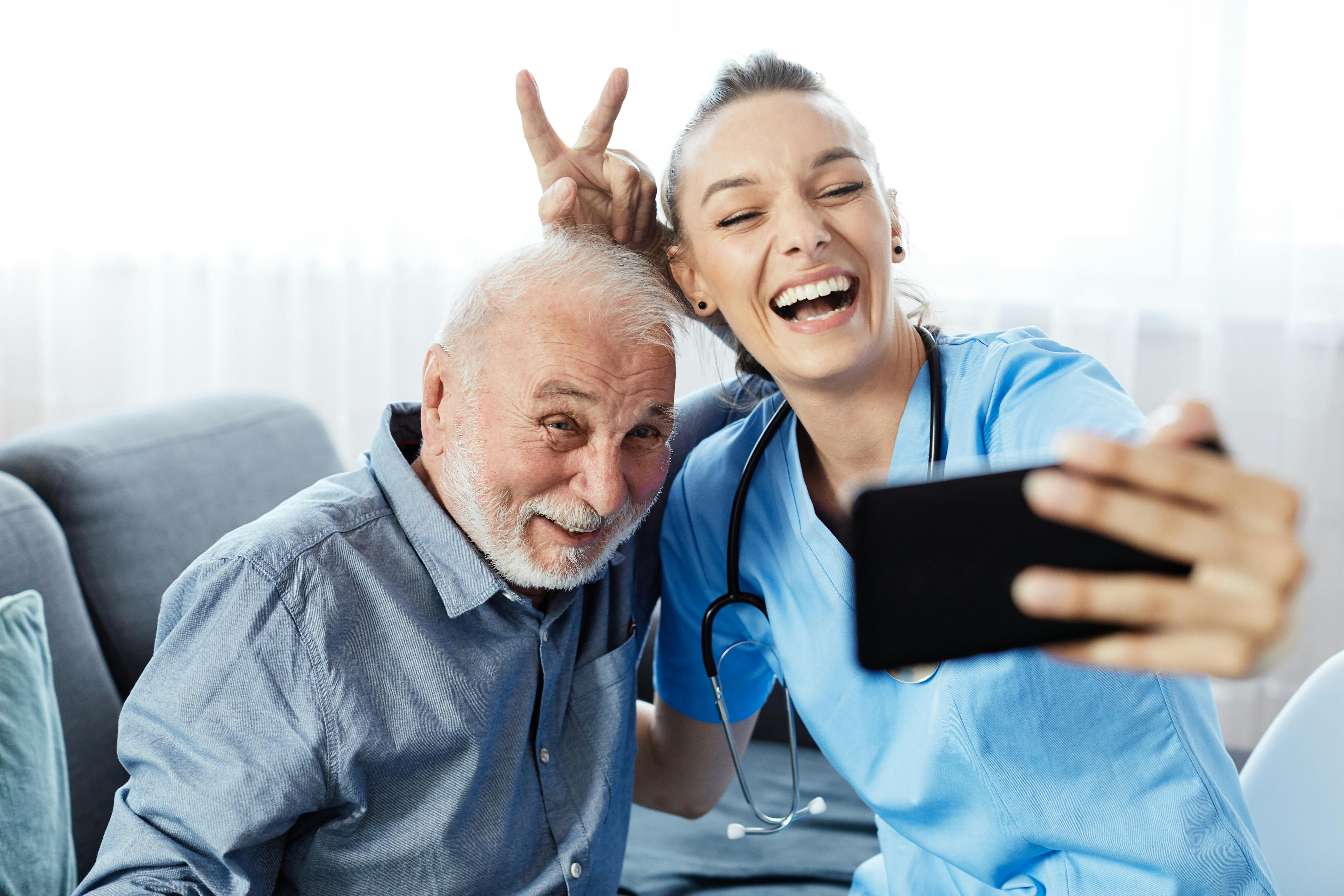 A young woman caretaker and an elderly man smile together while they pose for a selfie.