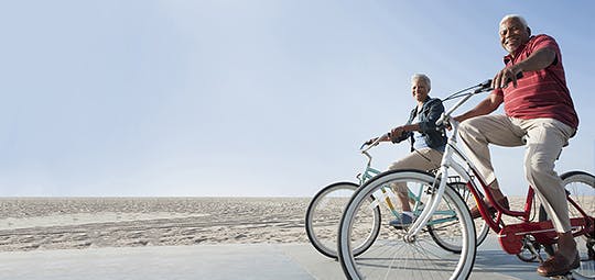Eldre kvinne og mann av afrikansk bakgrunn sykler langs en strand.
