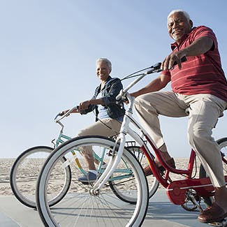 Eldre kvinne og mann av afrikansk bakgrunn sykler langs en strand.